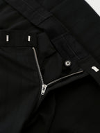 Close-up detail of black trousers showing a zippered fly, button closure, belt loops, and fabric texture.