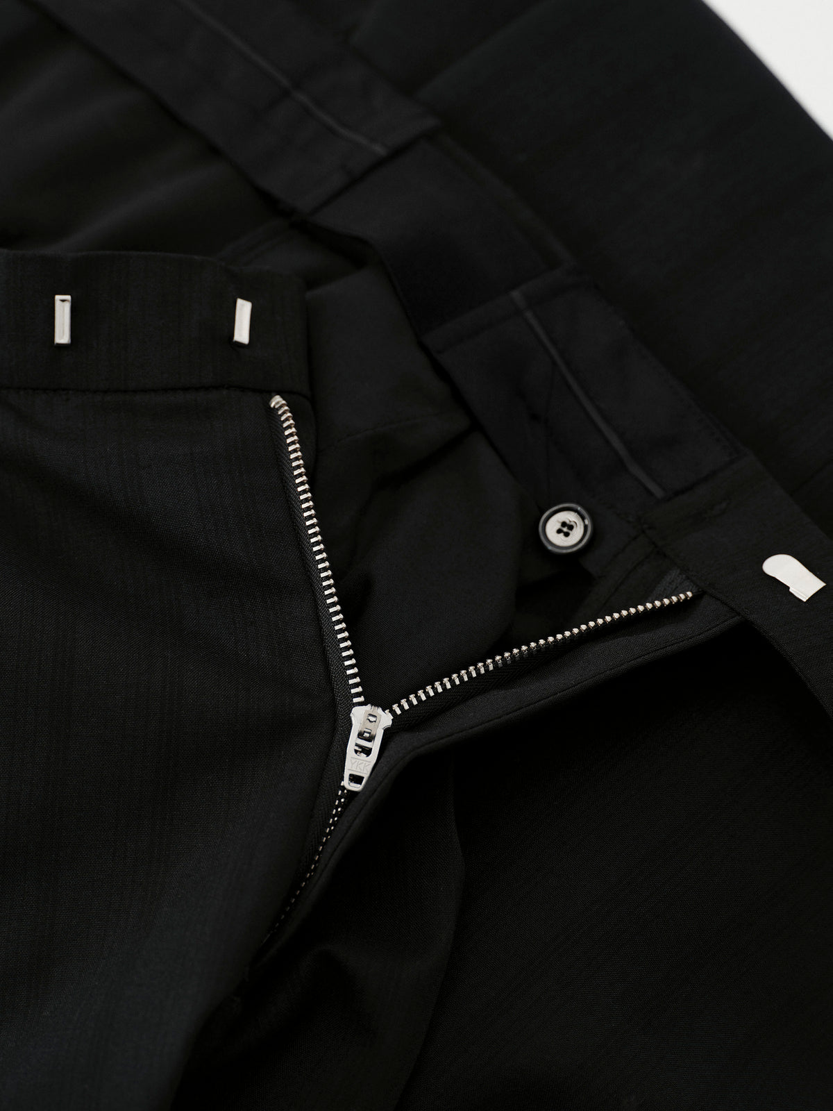Close-up detail of black trousers showing a zippered fly, button closure, belt loops, and fabric texture.