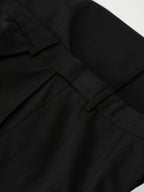 Close-up of black pleated fabric with a structured waistband and belt loops, photographed against a small white background.