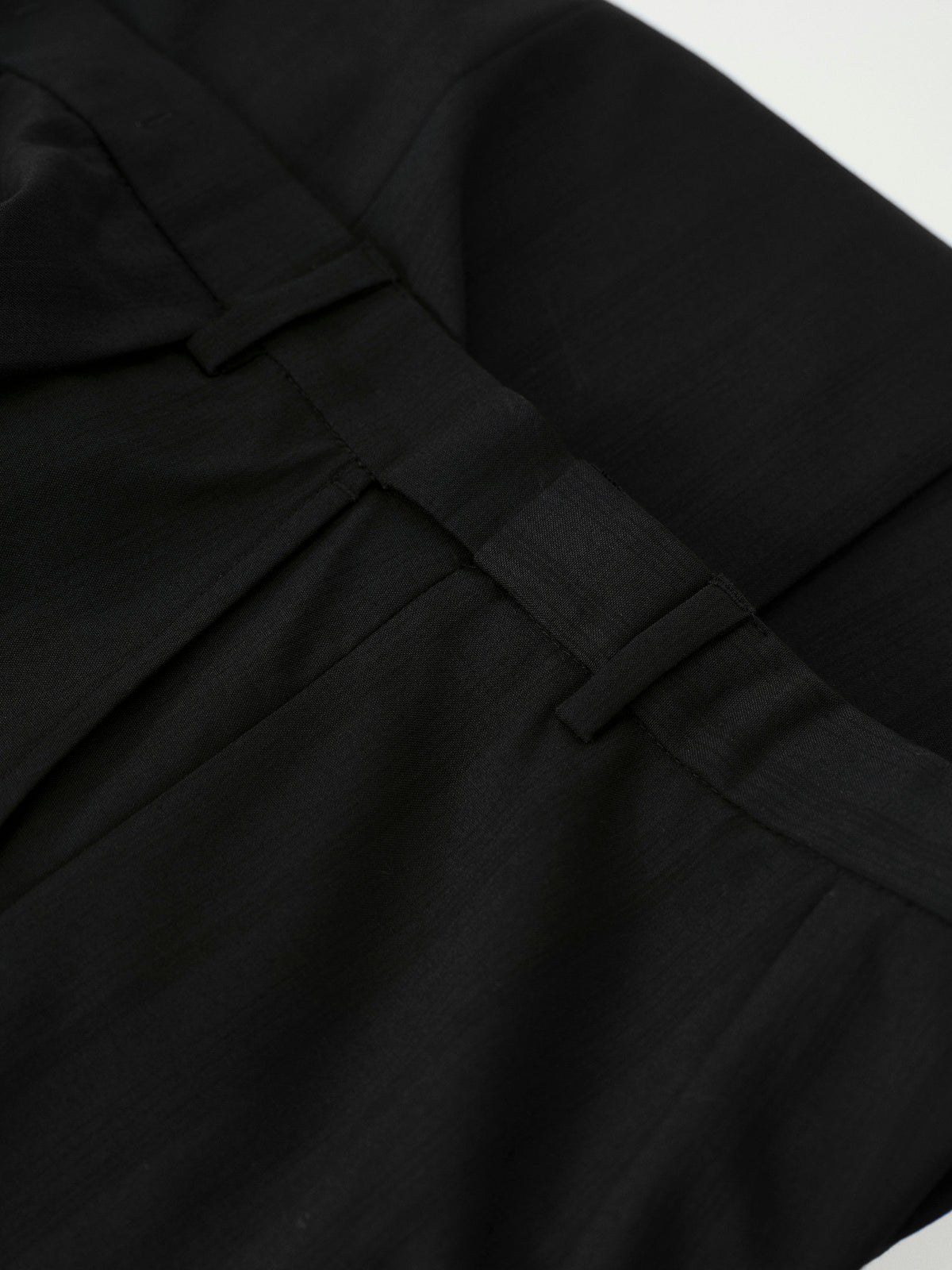 Close-up of black pleated fabric with a structured waistband and belt loops, photographed against a small white background.