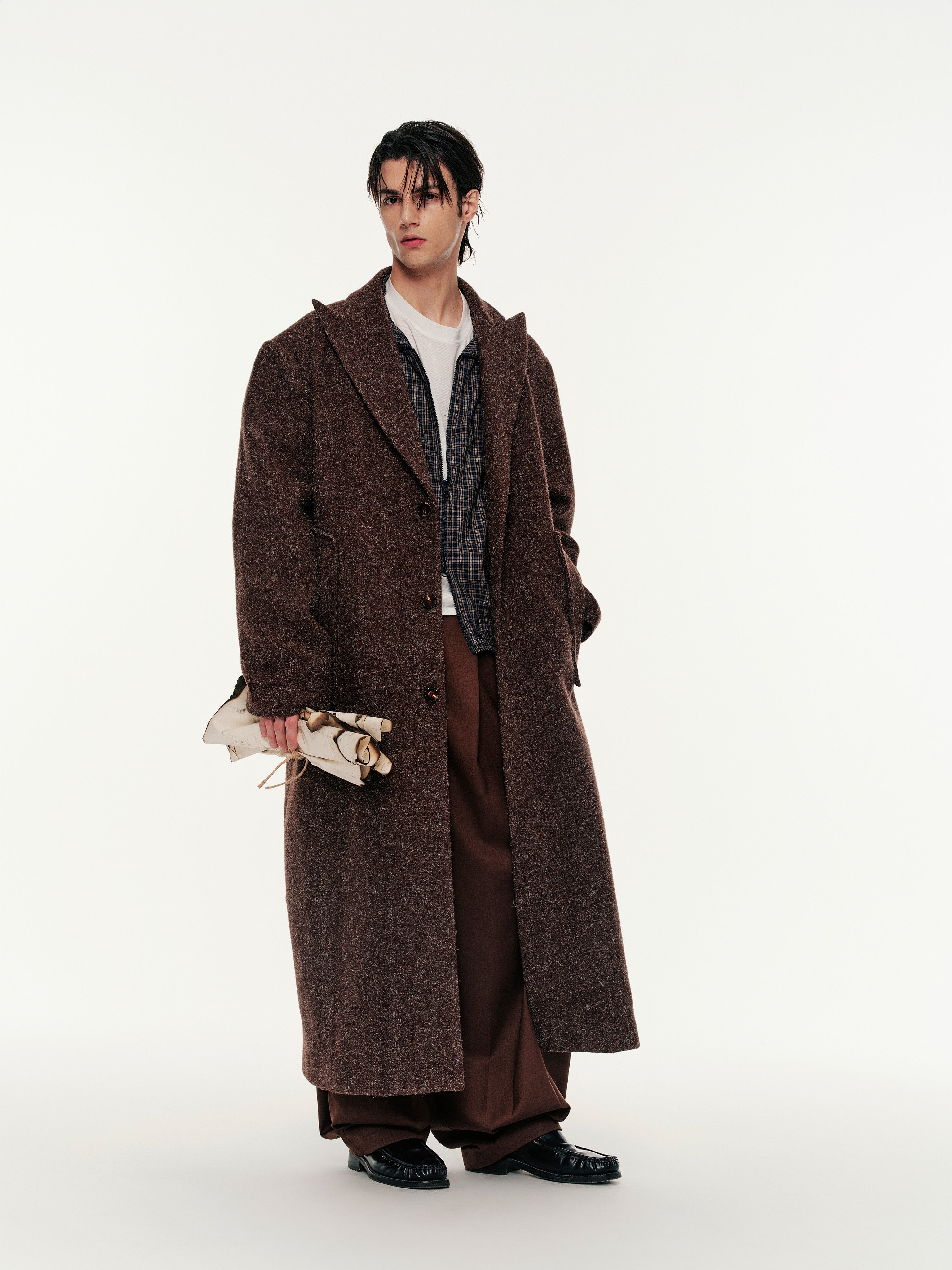 Man wearing a long brown coat holding a patterned bag on a white background