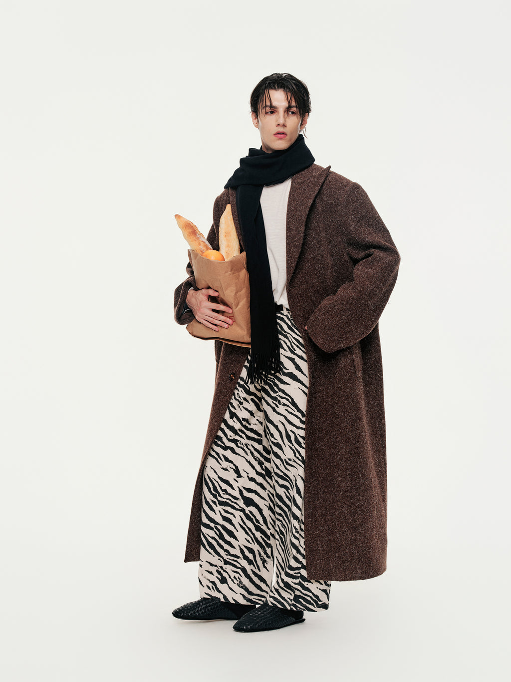 Person wearing a long brown coat and zebra print pants holding a paper bag on a white background